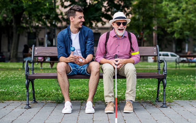 Alter blinder Mann auf einer Parkbank lacht zusammen mit einem jungen Mann